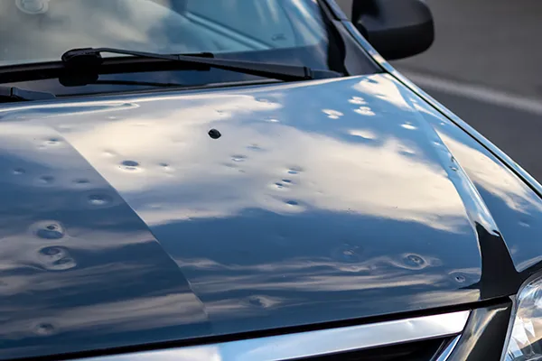 Close-up of a black car hood covered in numerous small dents caused by severe hail damage, highlighting the need for specialized paintless dent repair services.