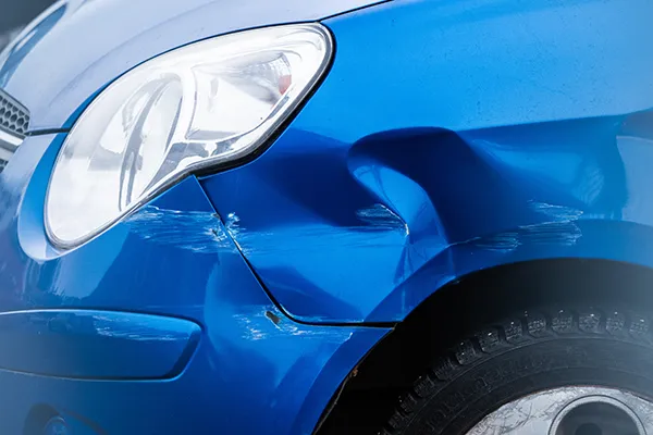 Close-up of a blue car showing a large dent and white paint transfer scratches on the front fender near the headlight, indicating minor collision damage.