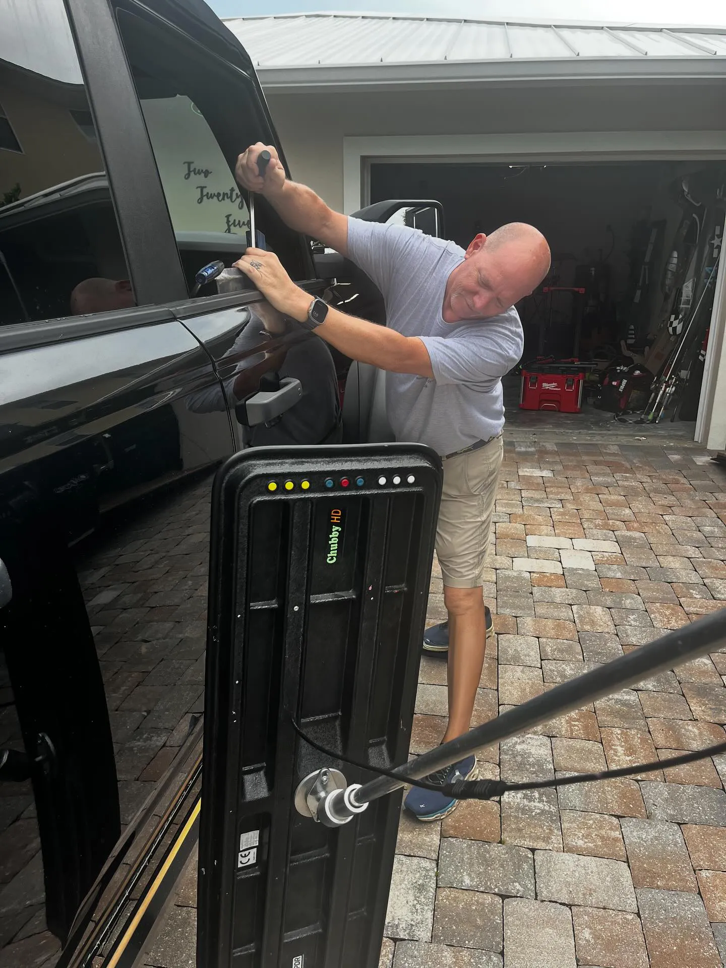 Professional PDR specialist Chris using a specialized metal rod tool to repair a dent on a black truck door through the window seal, with a reflection board in the foreground