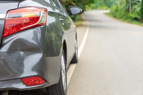 Close-up of a large dent on the rear bumper of a grey sedan parked on the side of a road, showing common collision damage requiring repair.