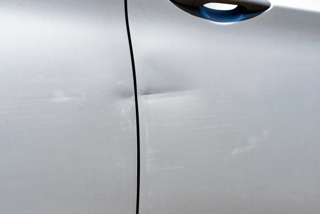 Close-up of a silver car door and side panel featuring a horizontal crease dent and surface scratches requiring paintless dent repair and detailing.