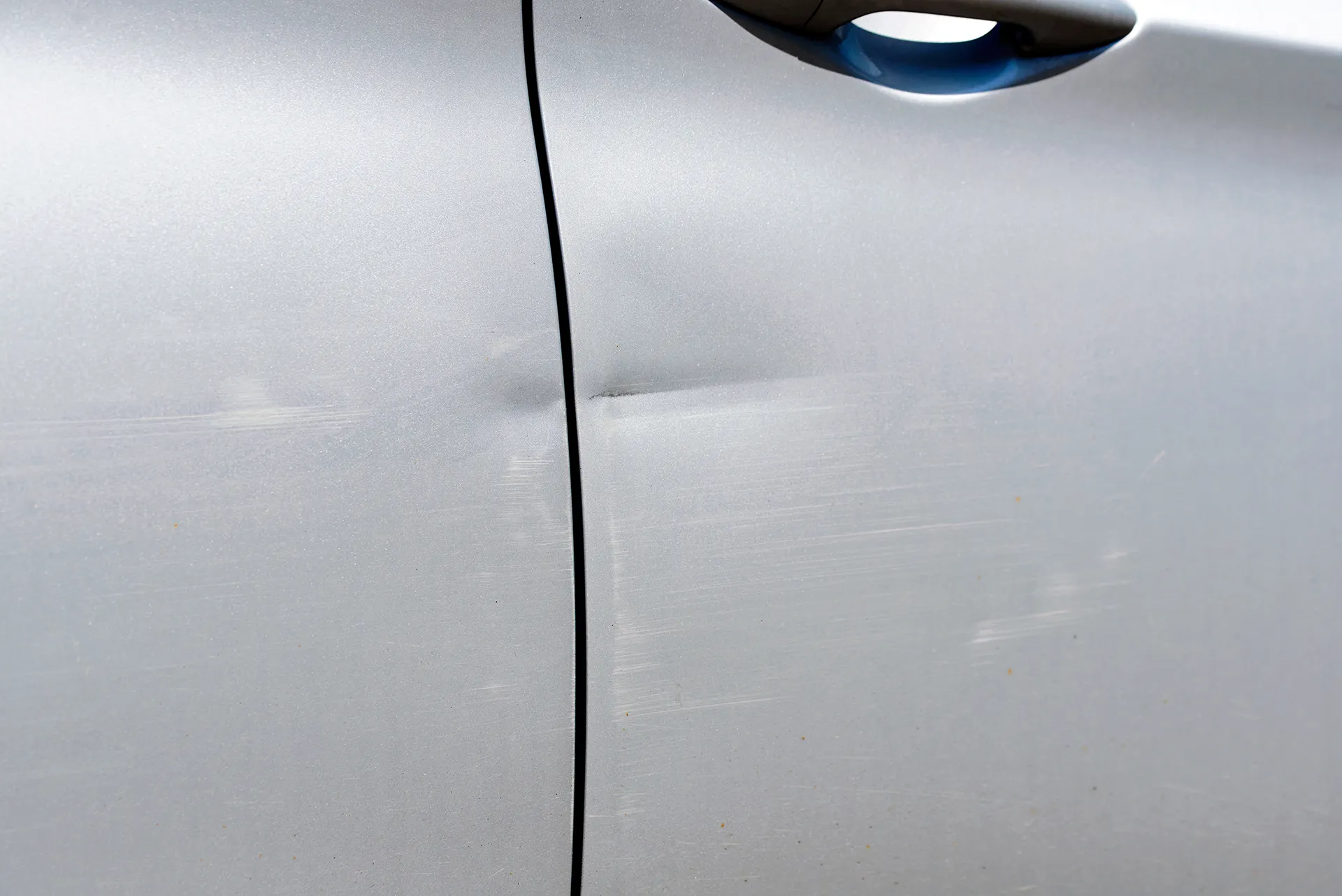 Close-up of a silver car door and side panel featuring a horizontal crease dent and surface scratches requiring paintless dent repair and detailing.